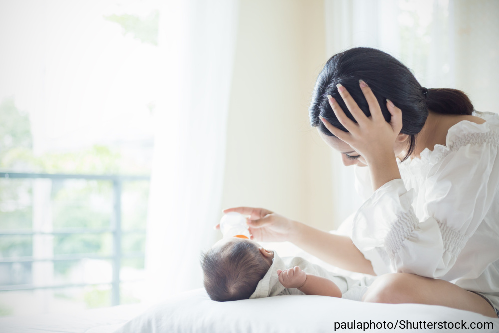 頭を抱えながら、赤ちゃんに哺乳瓶でミルクをあげているお母さん