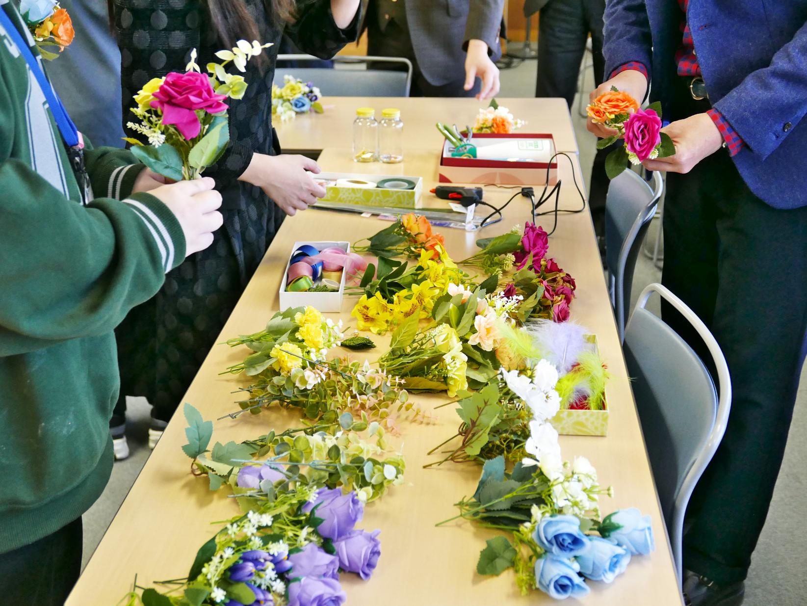 造花など材料が並びコサージュを作る人々