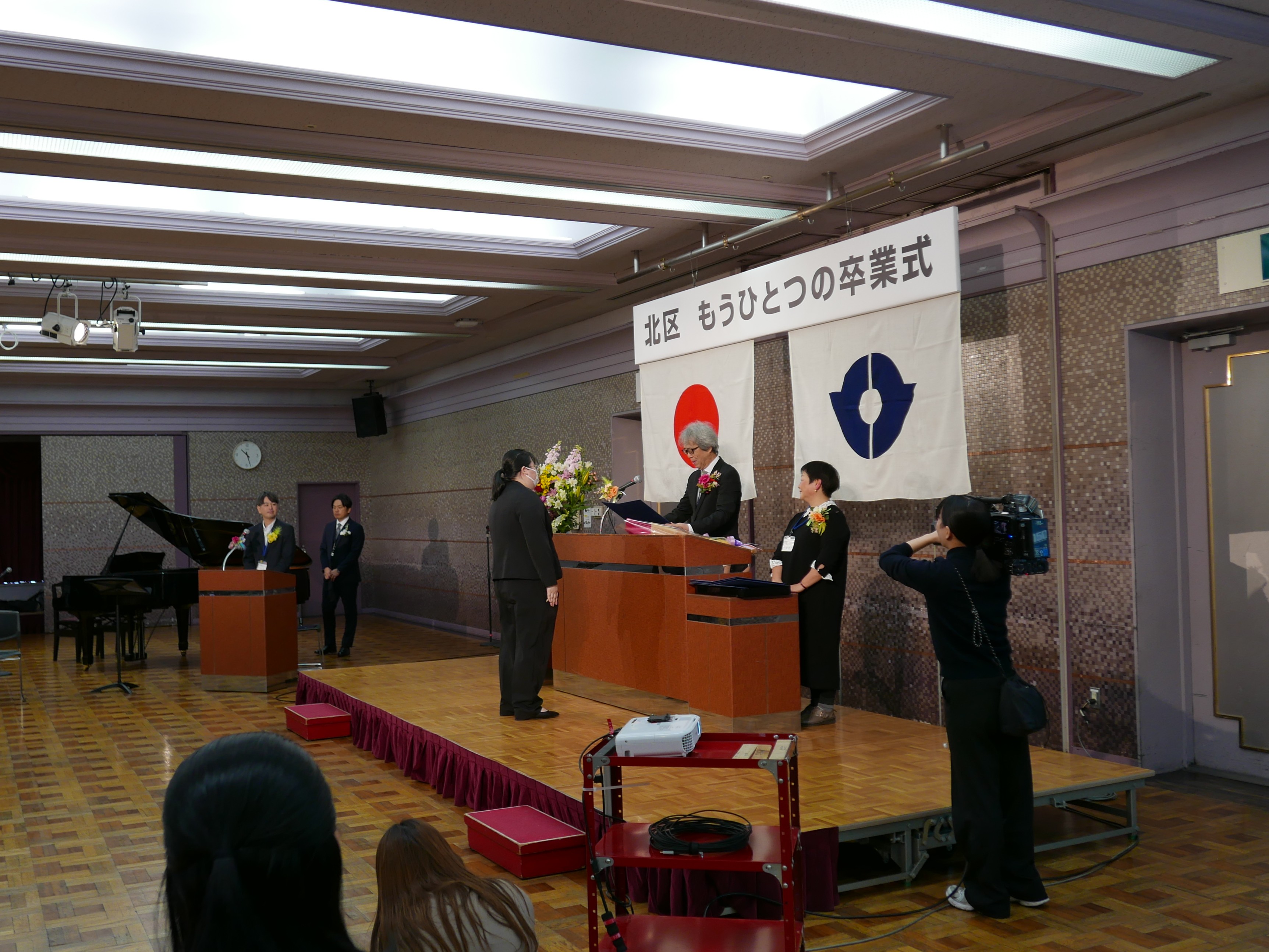 壇上に上がって、教育長から卒業証書を受け取る黒いスーツ姿の女性