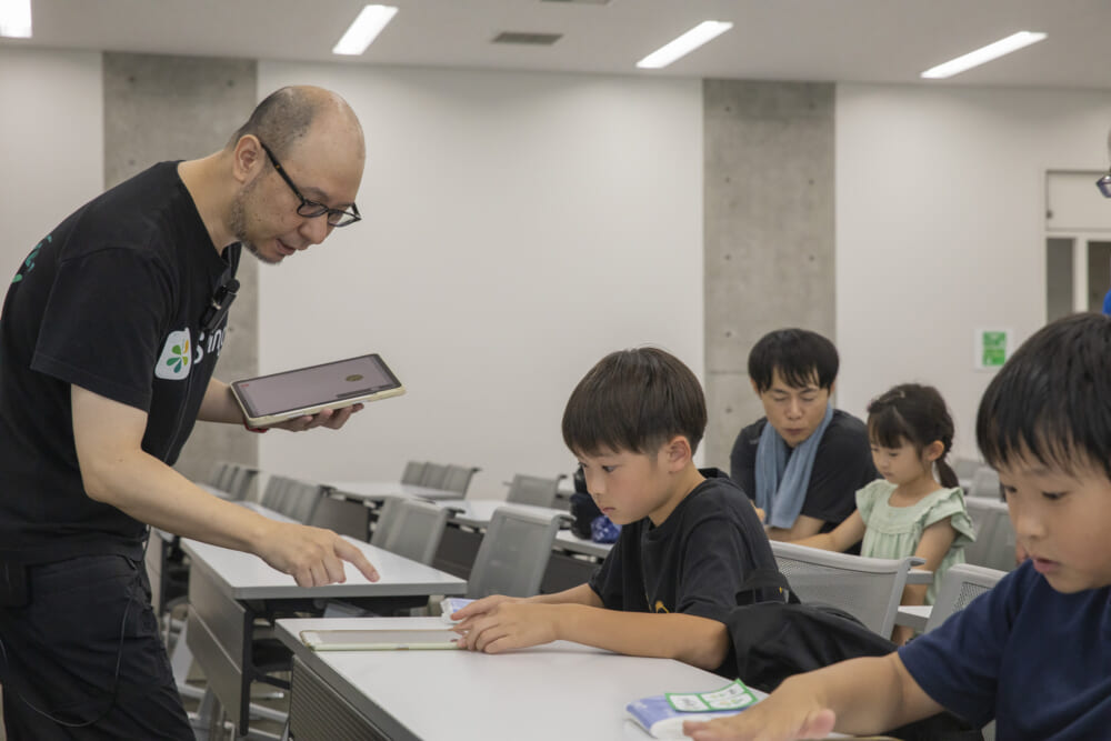先生が参加者の子どもたちの席に行って操作方法を教える様子