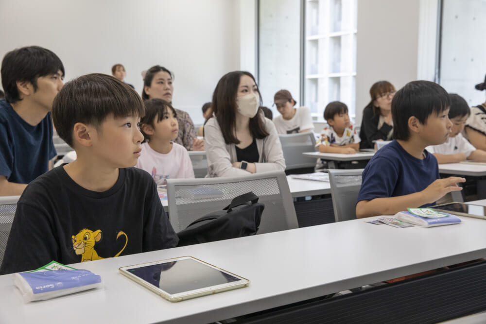 先生の操作画面が映った教室前のモニターを真剣に眺める参加者たち