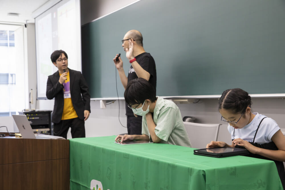 タブレットに集中してゲーム作りに励む2人と、その隣で実況と解説をする様子。
